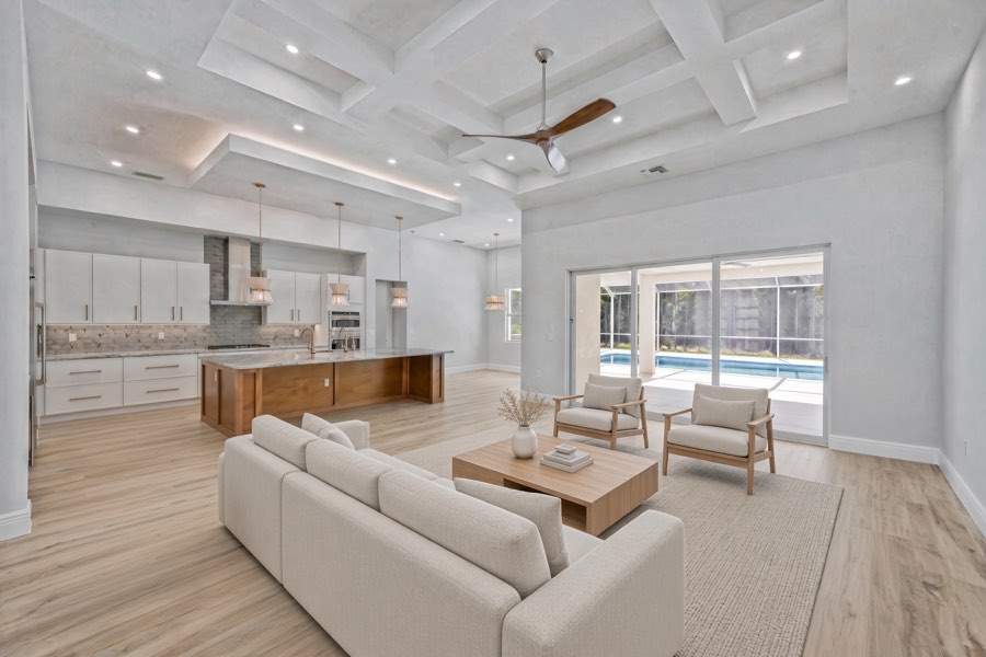 Bright open living room with a white sofa, coffered ceiling, kitchen island and sliding doors to the pool